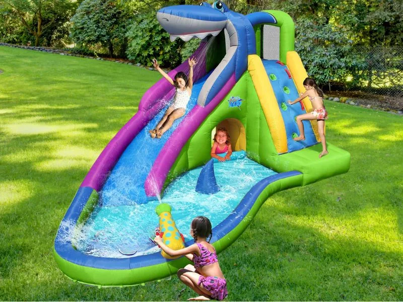 A girl sliding down the purple lane of an Action Air shark water slide into a curving pool on a lawn, other children climbing and splashing