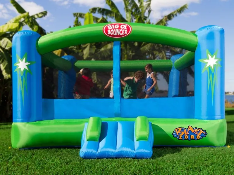 Children bouncing inside a Blast Zone Big Ol Bouncer with a green arch and blue corner pillars, on a lawn beside banana plants
