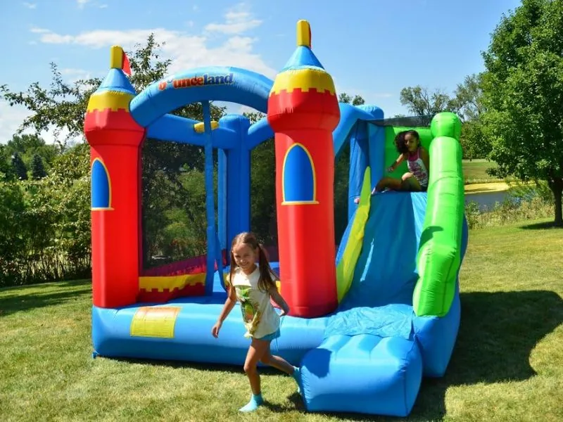 A girl walking past a Bounceland Royal Palace with red turrets on a sunny lawn while another child sits at the top of its green and blue slide