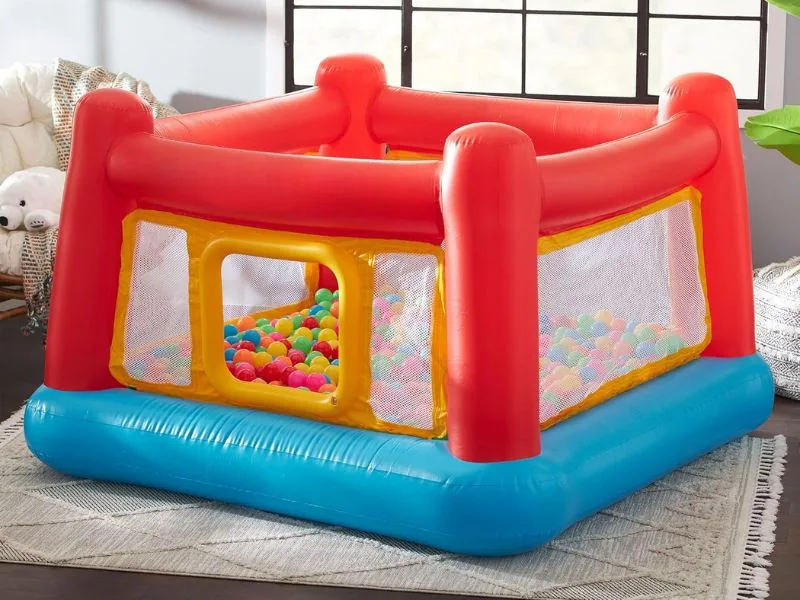 Intex Jump-O-Lene with red posts, a blue base and mesh sides, half full of pit balls on a living-room rug