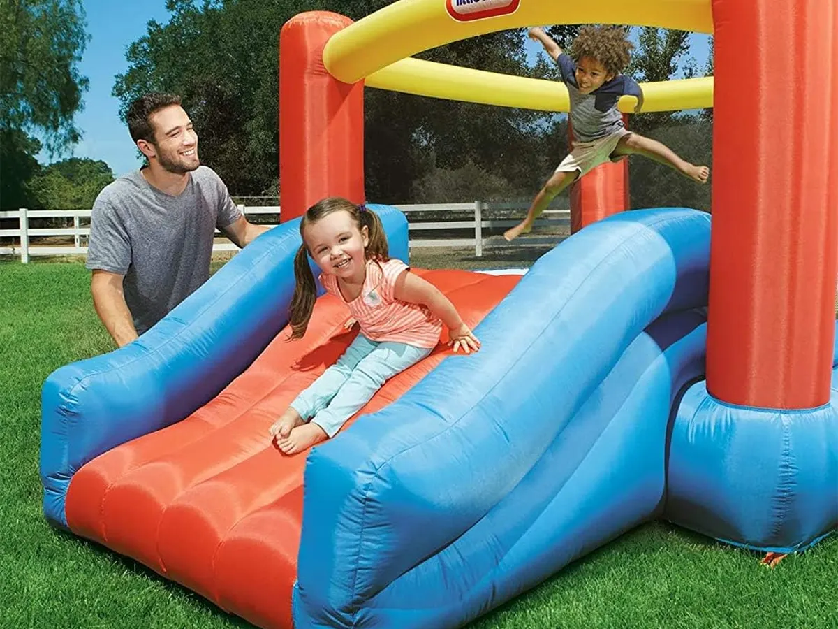 A child on the slide of a Little Tikes Jr. Jump 'n Slide in a fenced backyard, a parent beside her