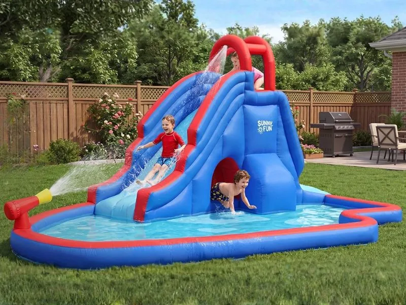 A child sliding down a Sunny & Fun Deluxe water park in blue and red while another crawls out of its tunnel into the splash pool, in a fenced backyard